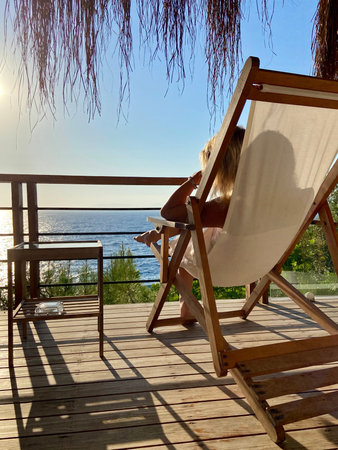Young caucasian woman sitting relaxed on a textile and wood sunbed watching the sea and sunset....