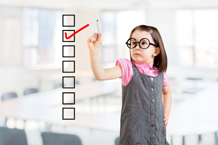 Cute little girl wearing business dress and checking on checklist box. Office background.の写真素材