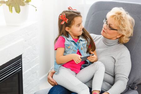 Grandmother and grandkid holding smartphone in hands, girl teaching grannyの写真素材