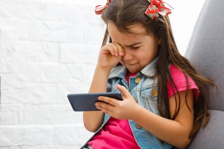 Little girl crying and holding smartphone.The little girl hid her face and cried, holding a mobile phone in her hands.の写真素材