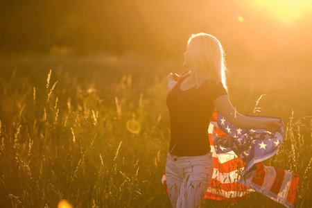 Beautiful young girl holding an American flag in the wind in a field of rye. Summer landscape against the blue sky. Horizontal orientation.の写真素材