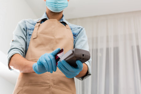 Cashier hand holding credit card reader machine and wearing disposable gloves, paying with smartphone during pandemic.の写真素材