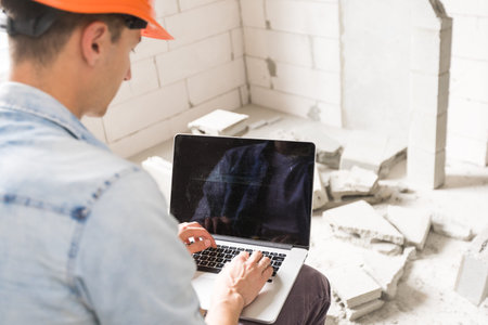 Handsome architect or foreman in helmet working with laptop on the apartment drawings at the...