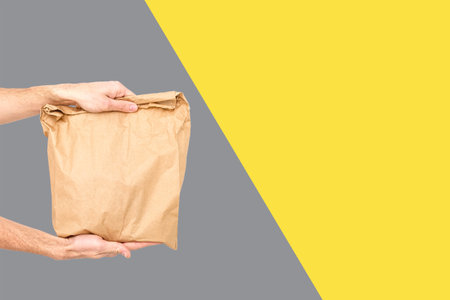 Close up female holds in hand brown clear empty blank craft paper bag for takeaway Delivery service concept. Copy space. Trendy colors 2021 - Gray and Yellow.の写真素材