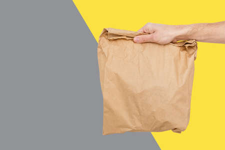 Close up female holds in hand brown clear empty blank craft paper bag for takeaway Delivery service concept. Copy space. Trendy colors 2021 - Gray and Yellow.の写真素材