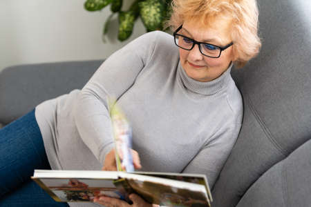 Cheerful senior female holding family photo album sitting on sofa at home, elderly woman watching photobookの写真素材