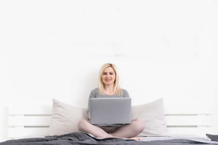 Smiling blonde woman using laptop at home in bedroom. Working from home in quarantine lockdown. Social distancing Self Isolationの写真素材