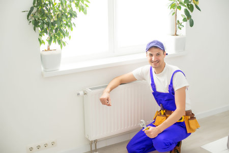 Man in work overalls using wrench while installing heating radiator in room. Young plumber...