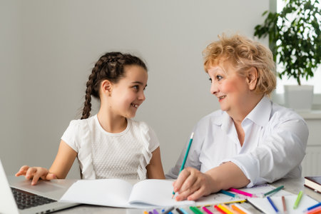 Happy grandchild with grandmother having fun, drawing colored pencils, sitting together at home, laughing preschool girl with smiling grandma painting picture, playing with granddaughterの写真素材