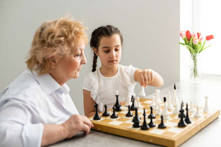 Happy girl with grandmother sitting at the table and playing chessの写真素材