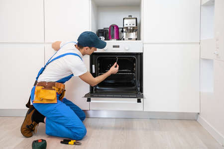 young repairman in protective workwear fixing oven in kitchenの写真素材