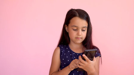 Little preschooler girl in a dress holding a mobile phone, isolated over pink