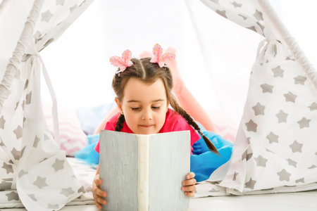Enjoying great story. Cute little girl reading book while sitting on the floor in tent