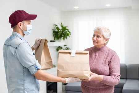 Young male volunteer in mask gives an elderly woman boxes with food near her house. Son man helps a...