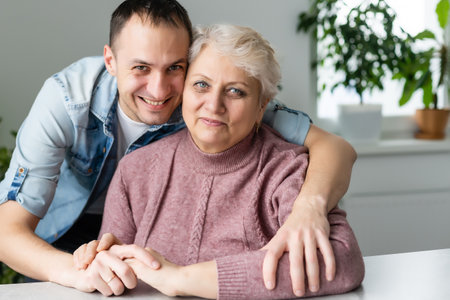 Head shot happy young bearded man embracing beautiful smiling middle aged senior mother in eyewear, relaxing together on comfortable sofa in living room, enjoying funny conversation or gossipingの写真素材