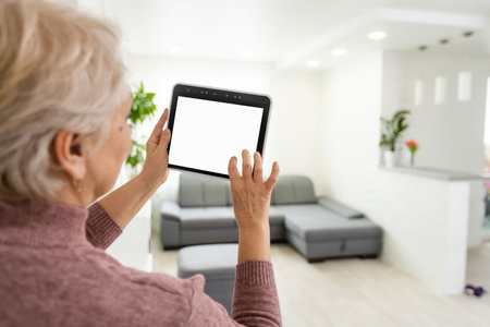 Senior woman at home standing at kitchen holding digital tablet controlling smart home system back view checking cameras close-upの写真素材