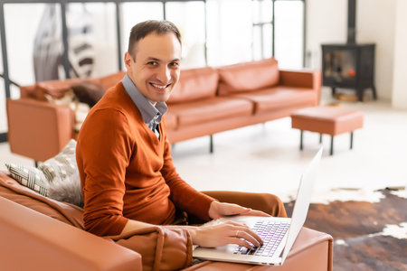 Young man smiling, reads the screen of a laptop computer while relaxing on a comfortable couch at...