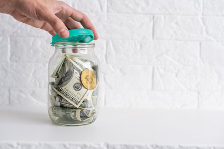 Saving money coin in jar. Symbol of investing, keeping money concept. Collecting cash banknotes in glass tin as moneybox. American dollars close-up with increasing columns of gold coins on tableの写真素材