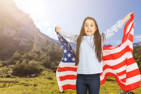 Young little girl holding American flag on amazing sky, mountain and meadow nature background at...