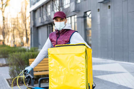 Young courier in medical mask delivering food with yellow thermal backpack, riding a bicycle in the city. Food delivery service conceptの写真素材