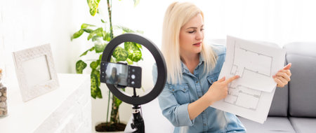 Calm clever woman standing in the bedroom in front of a camera on tripod and holding a roll of paper