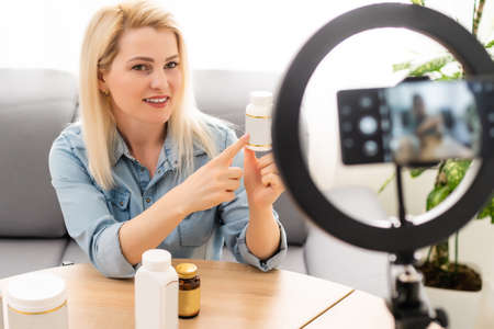 blogger woman recording video on smartphone and lighting herself with ring lamp at table in light roomの写真素材
