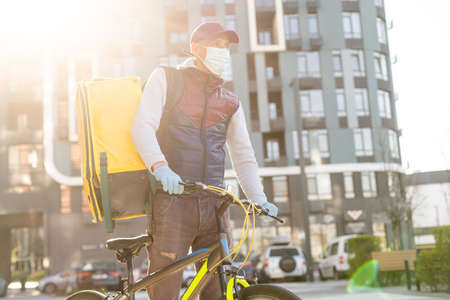 Young courier in medical mask delivering food with yellow thermal backpack, riding a bicycle in the city. Food delivery service conceptの写真素材