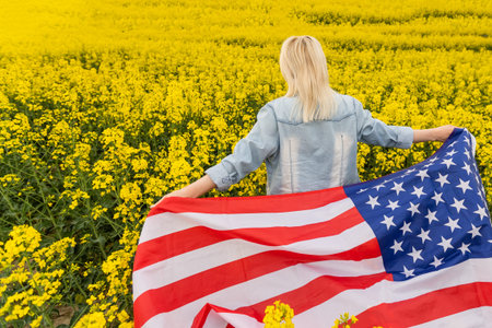 attractive woman holding an American flag in the wind in a field of rapeseed. Summer landscape...