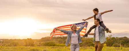 Young parents with their daughter holding American flag in countryside at sunset. Independence Day celebrationの写真素材