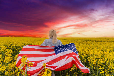 Adult woman holding American flag with pole, stars and stripe in a yellow rapeseed field. USA flag fluttering in the windの写真素材