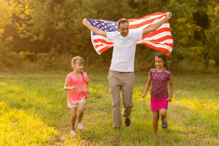 Patriotic holiday. Happy family, mother and daughters with American flag outdoors on sunset. USA celebrate independence day 4th of July.の写真素材
