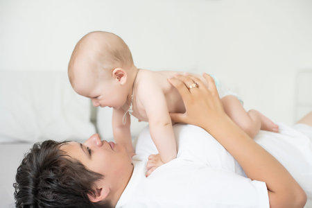 Mother and child on a white bed. Mom and baby boy in diaper playing in bedroom. Parent and little...