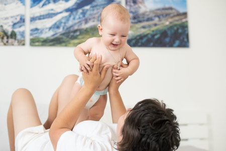 Mother and child on a white bed. Mom and baby boy in diaper playing in bedroom. Parent and little...