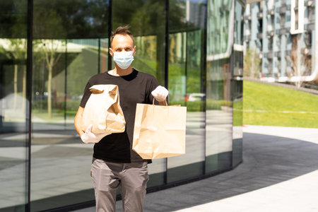 Delivery guy with protective mask and gloves holding bag with groceries in front of a building.