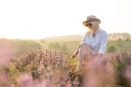 Beautiful young healthy woman with a white dress running joyfully through a lavender field holding a straw hat under the rays of the setting sunの写真素材