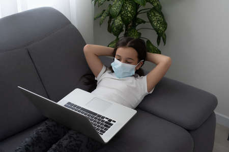 Smart Little Girl wearing mask Does Homework in Her Living Room. Shes Sitting and Uses Laptop.の写真素材