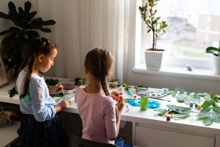 Cute schoolgirl with scissors cutting dry oak leaf while helping her teacher with decorations for holiday at lessonの写真素材