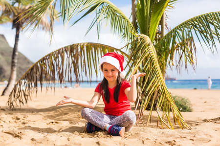 Little girl in santa hat sitting next to palm tree on beachの写真素材