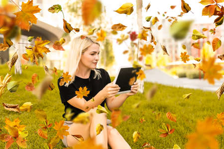 woman sitting with a tablet in her hands fall city park on a warm day. Autumn golden leavesの写真素材
