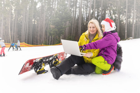 mother and daughter with a snowboard using laptop on snowy mountainの写真素材
