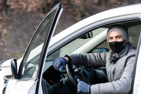 Horizontal shot of man wears spectacles and protective medical face mask, stands near car, commutes to work by personal transport during quarantine. Travel and pandemic situation.の写真素材