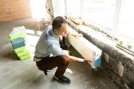 Full length shot of professional plumber, worker in uniform fixing or installing heating radiator. Construction, maintenance and repair concept. Selective focus. Horizontal shotの写真素材