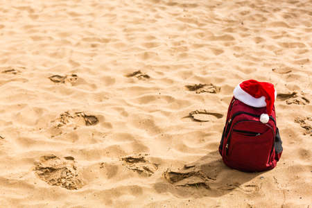 Santa hat on a tourist red backpack against the blue sea, enjoying the sunset of the ocean horizon. Christmas beach vacation travel banner or cardの写真素材