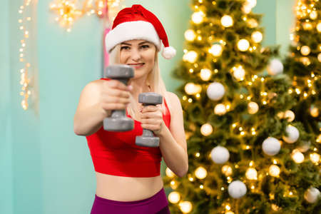 Festive fit blonde holding dumbbell against blurry christmas tree in roomの写真素材