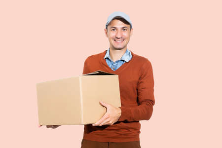 Cheerful delivery man. Happy young courier holding a cardboard box and smiling while standing against pink backgroundの写真素材