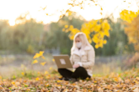 Life during covid-19. Young woman wearing protective face mask typing on laptop in a public park. Stylish female using computer while working outdors. Distance learning and remote work during pandemicの写真素材