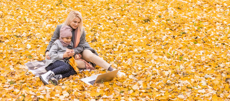 Mom and daughter resting on nature with laptop sitting on an autumn dayの写真素材