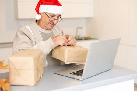 Senior man grandfather in Santa festive hat sitting on sofa and communicating with family online during videocall on laptop at Christmas decorated home. Pensioner congratulating children online.の写真素材