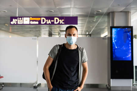 Young European man in gray coat, protective disposable medical mask in airport. Afraid of dangerous N-CoV 2019 influenza coronavirus. backpack, suitcase.の写真素材