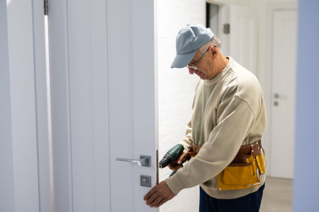 Attractive builder is installing lock in door. He is holding a screwdriver and kneeling.